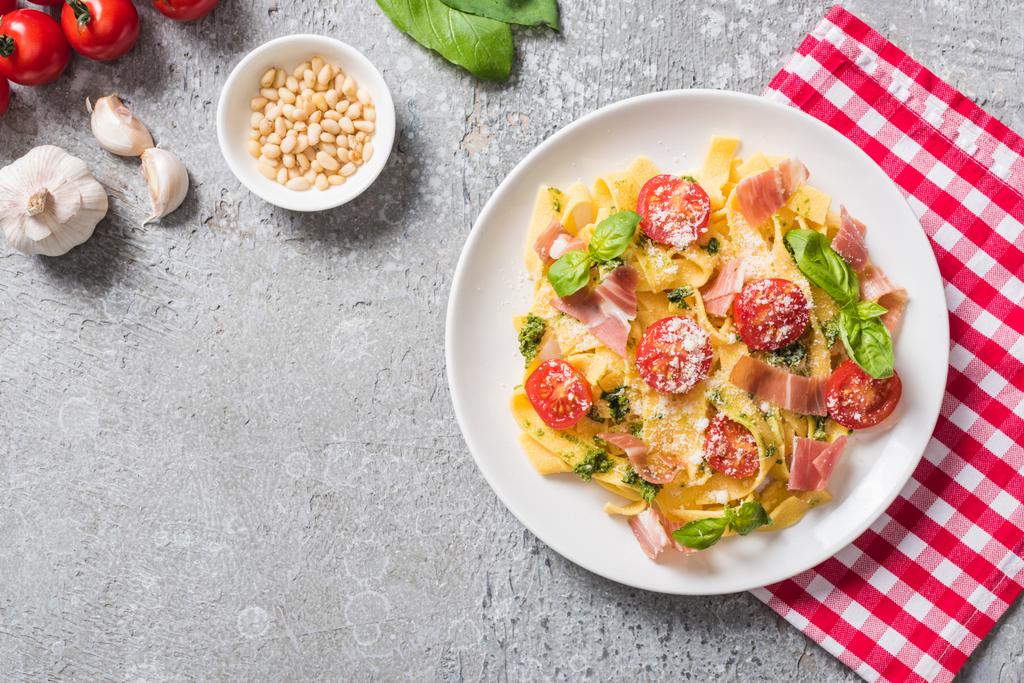 Draufsicht auf gekochte Pappardelle mit Tomaten, Basilikum und Prosciutto auf karierter Serviette nahe Zutaten auf grauer Oberfläche - Foto, Bild