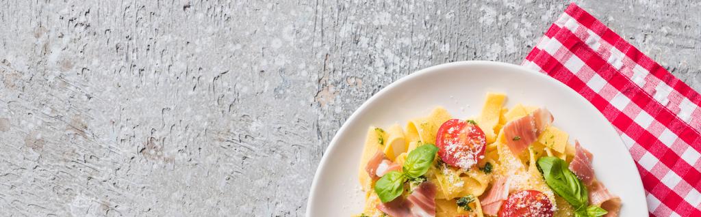 Draufsicht auf gekochte Pappardelle mit Tomaten, Basilikum und Schinken auf karierter Serviette auf grauer Oberfläche, Panoramaaufnahme - Foto, Bild