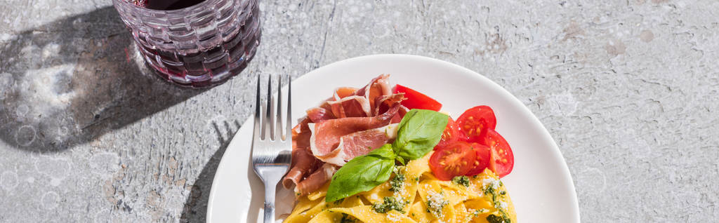 widok z góry smaczne Pappardelle z pomidorami, pesto i prosciutto z widelcem w pobliżu czerwonego wina na szarej powierzchni, panoramiczny strzał - Zdjęcie, obraz