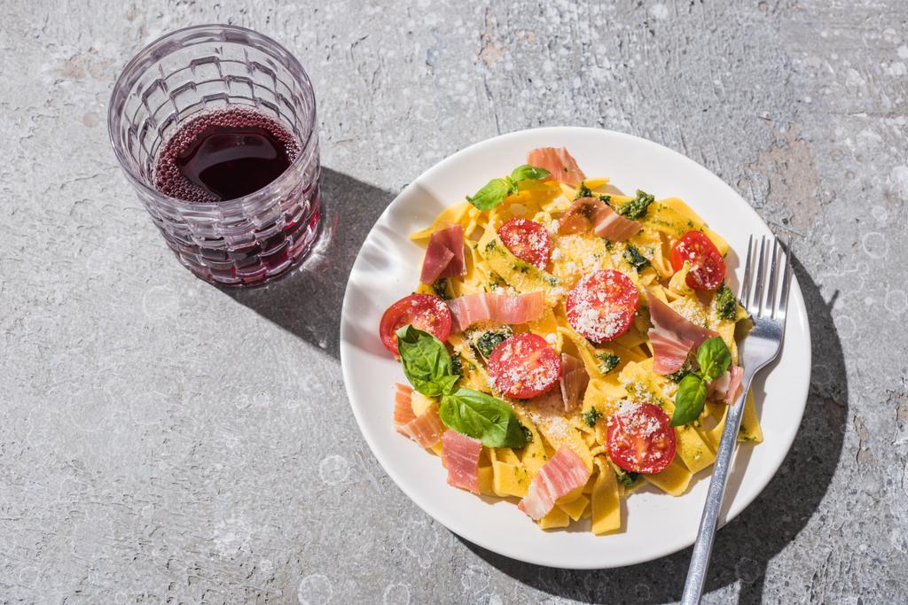 top view of tasty Pappardelle with tomatoes, pesto and prosciutto with fork near red wine on grey surface - Photo, Image