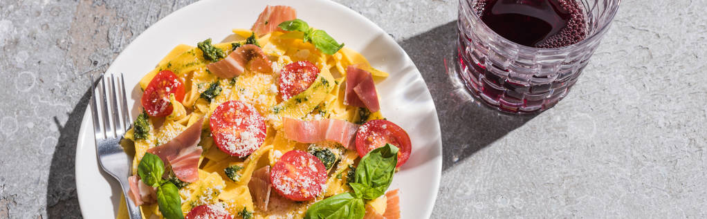 top view of tasty Pappardelle with tomatoes, pesto and prosciutto with fork near red wine on grey surface, panoramic shot - Photo, Image