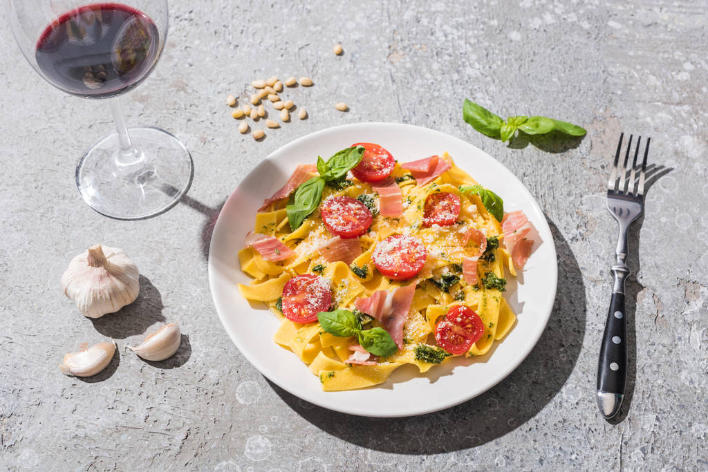 Pappardelle com tomates, pesto e prosciutto perto de vinho tinto e ingredientes na superfície cinza
 - Foto, Imagem