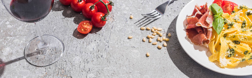 frische Pappardelle mit Tomaten, Pesto und Prosciutto in der Nähe von Rotwein und Zutaten auf grauer Oberfläche, Panoramaaufnahme - Foto, Bild