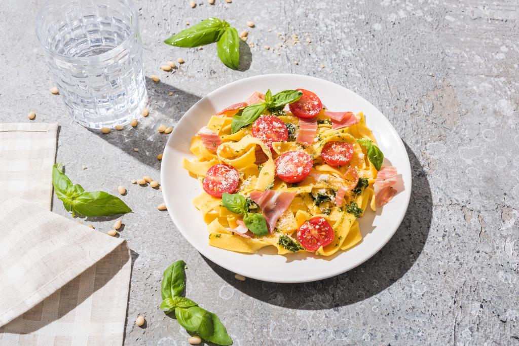 saboroso Pappardelle com tomates, pesto e prosciutto perto de água, guardanapo e pinhões na superfície cinza
 - Foto, Imagem