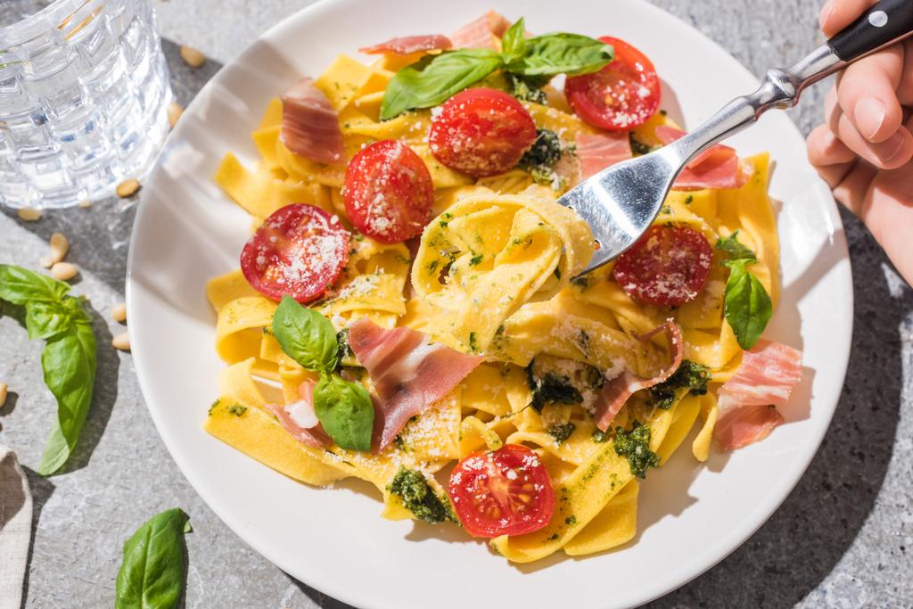 partial view of woman eating tasty Pappardelle with tomatoes, pesto and prosciutto near water on grey surface - Photo, Image