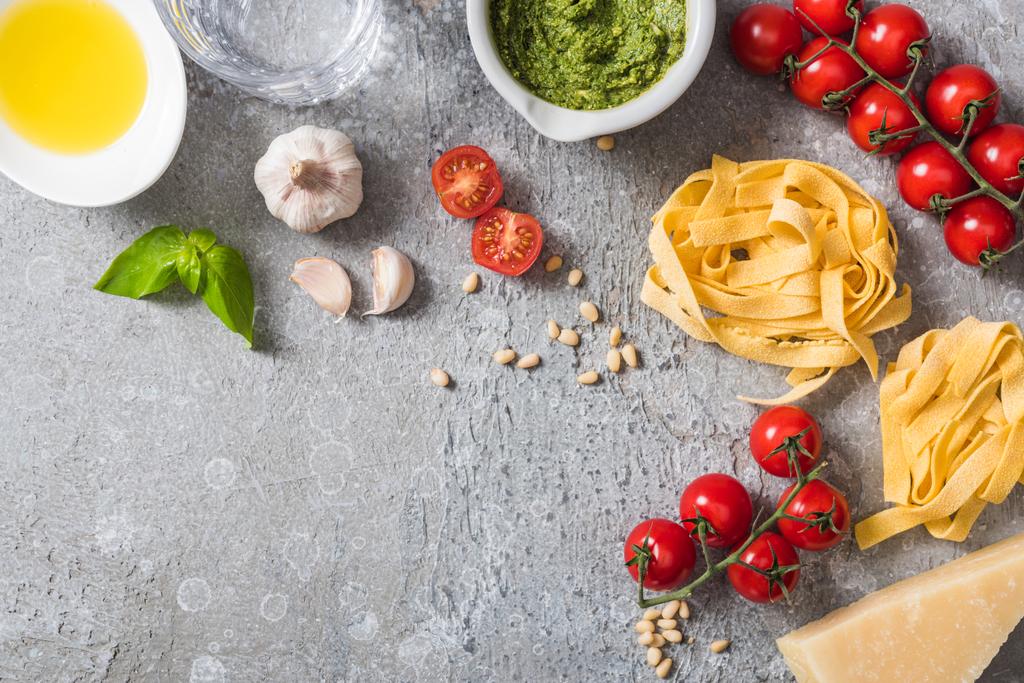 Blick von oben auf rohe Pappardelle in der Nähe von Tomaten, Knoblauch, Basilikum, Pinienkernen, Olivenöl, Wasser und Pesto-Sauce auf grauer Oberfläche - Foto, Bild