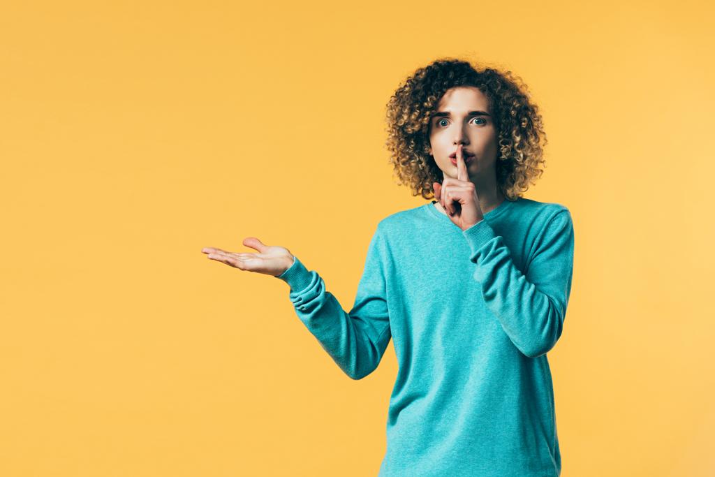 curly teenager showing shh gesture and pointing with hand aside isolated on yellow - Photo, Image