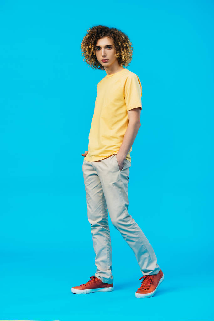 Вид в полный рост кудрявого подростка с руками в карманах, изолированных на голубом фоне
 - Фото, изображение
