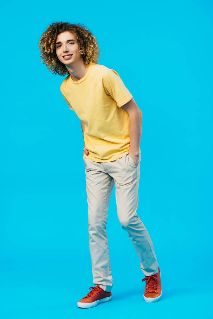 smiling curly teenager with hands in pockets isolated on blue - Photo, Image