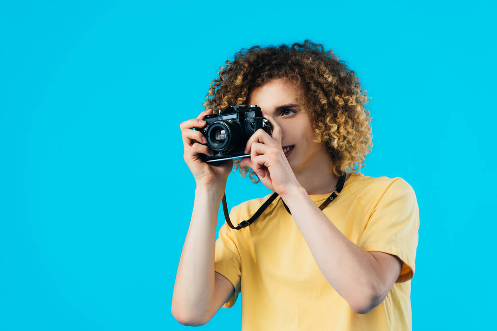 kudrnatý teenager fotografování na filmové kamery izolované na modré - Fotografie, Obrázek