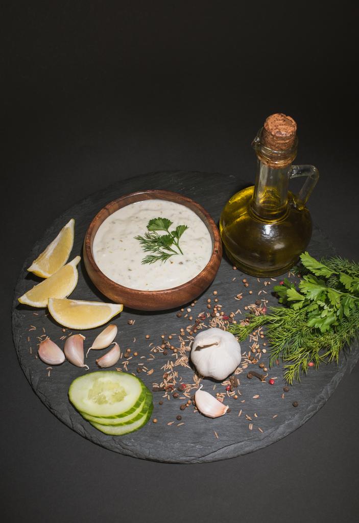 Zelfgemaakte tzatziki saus met ingrediënten, specerijen en olijfolie op donkere boord op zwarte achtergrond - Foto, afbeelding