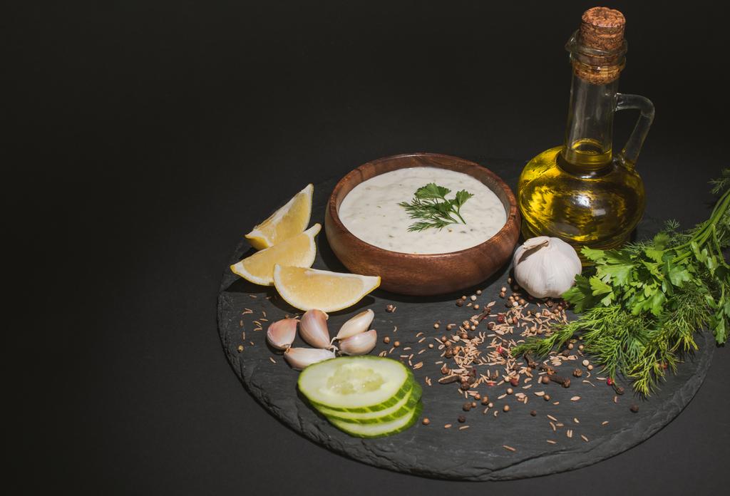Tzatziki saus met biologische ingrediënten, specerijen en olijfolie op donker bord op zwarte achtergrond - Foto, afbeelding