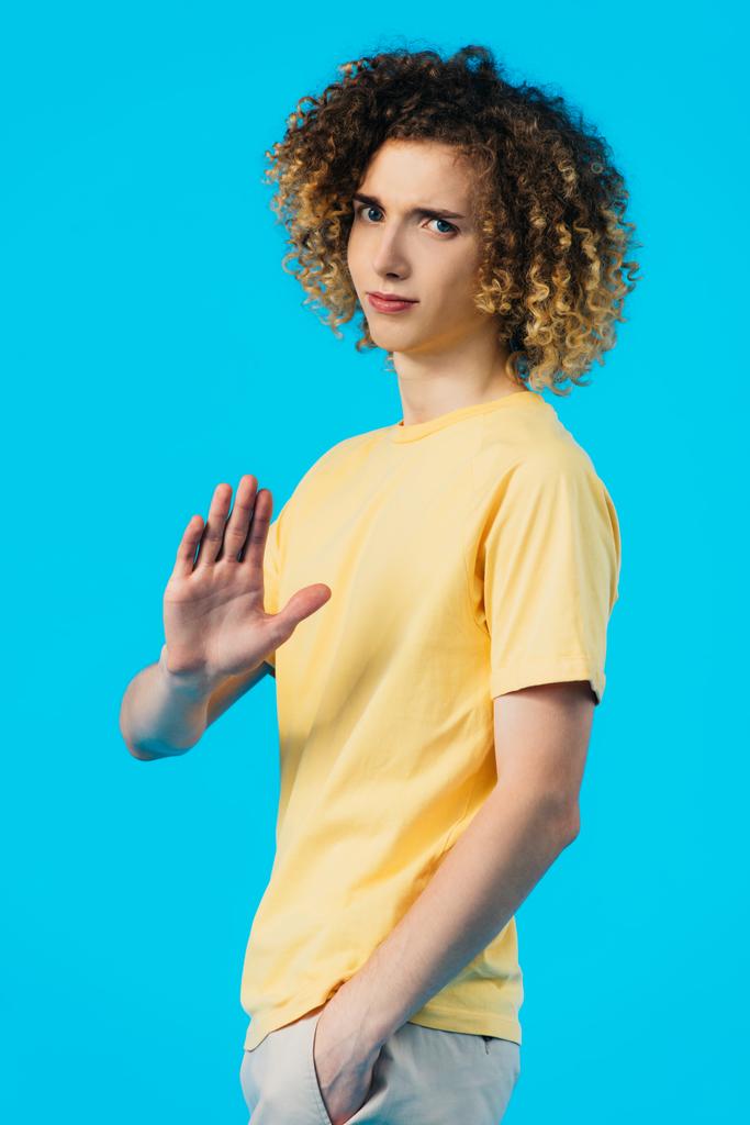 aufgebracht lockiger Teenager mit der Hand in der Tasche zeigt Stop-Geste isoliert auf blau - Foto, Bild