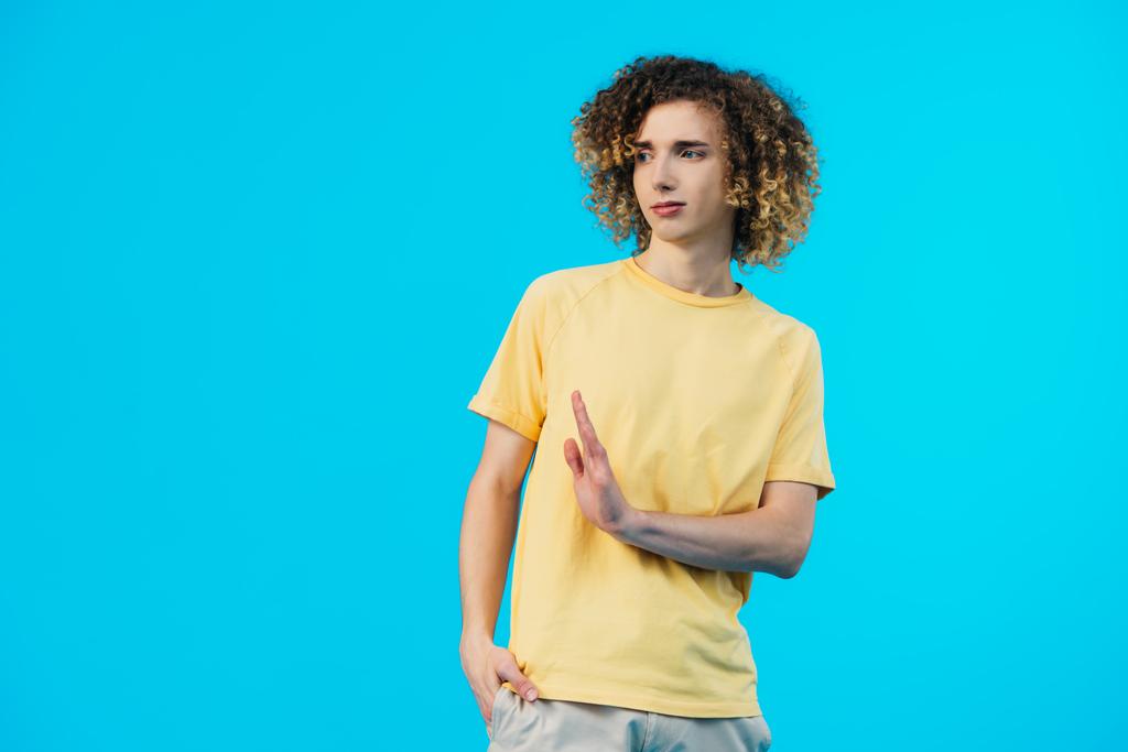 zmatený kudrnatý teenager s rukou v kapse ukazující stop gesto izolované na modré - Fotografie, Obrázek