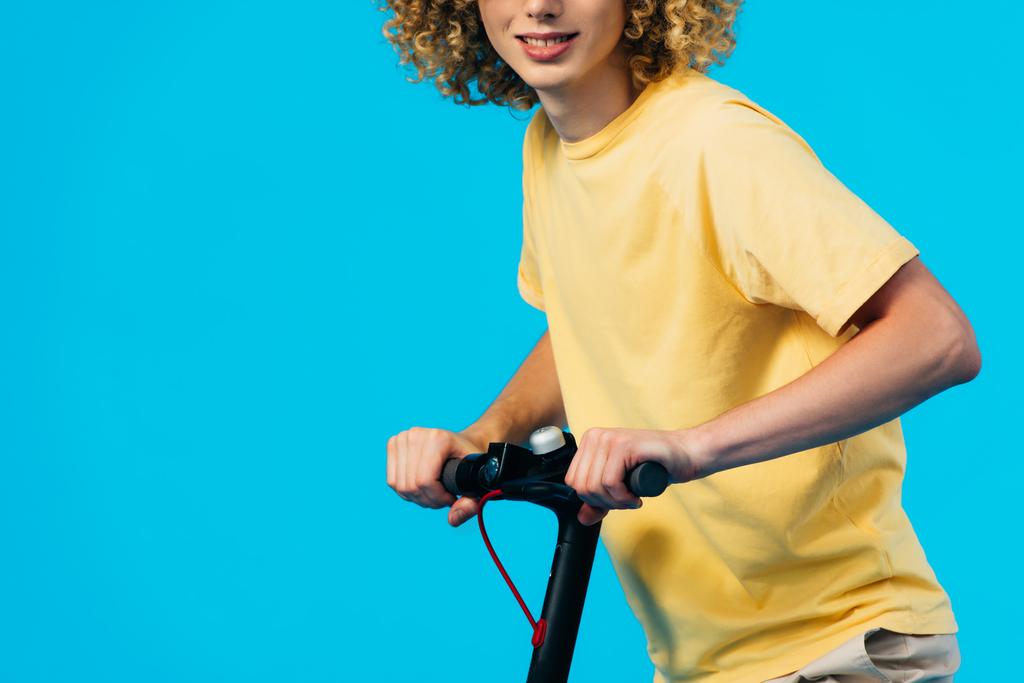vágott kilátás mosolygós göndör tinédzser lovaglás elektromos robogó elszigetelt kék - Fotó, kép