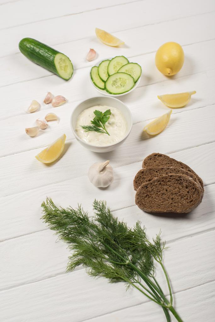Házi készítésű tzatziki szósz hozzávalókkal, zöldekkel és kenyérrel fehér fa alapon - Fotó, kép