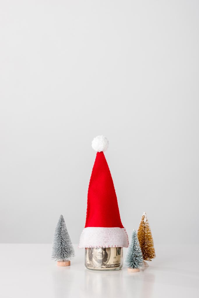 Dekoratif ve küçük çamlar kavanozun yanında dolar banknotları ve beyaz şapkalı Noel Baba.  - Fotoğraf, Görsel