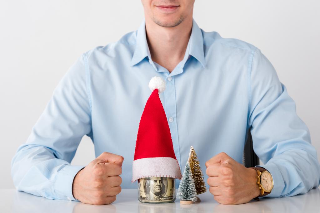 vista ritagliata di uomo con pugni serrati vicino vaso di vetro con soldi e cappello di Babbo Natale isolato su bianco
  - Foto, immagini