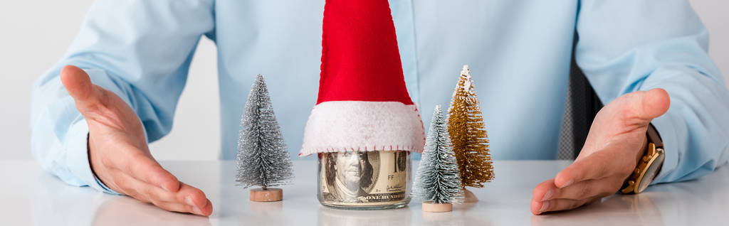 panoramic shot of man with pointing with hands at glass jar with money and santa hat isolated on white  - Photo, Image
