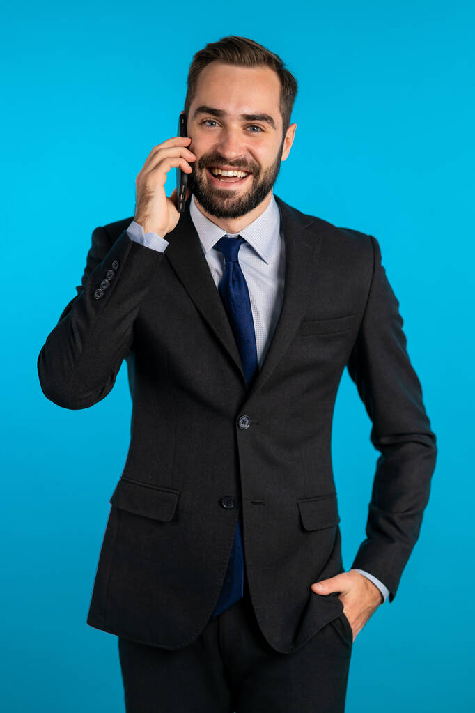 Бизнесмен беседует с помощью мобильного телефона, изолированного на синем фоне. Деловой парень в костюме с радостью беседует с коллегой. Работник офиса, наемный работник, концепция будней
 - Фото, изображение