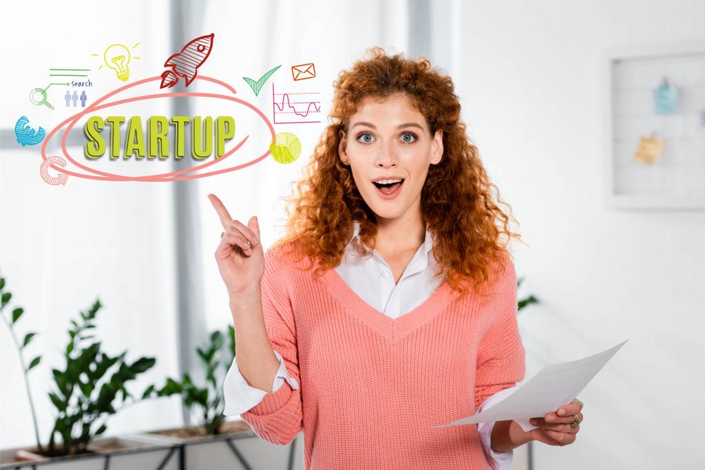 attractive and shocked businesswoman showing idea sign and holding paper with startup illustration - Photo, Image