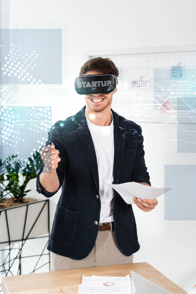 улыбающийся бизнесмен в рубашке с гарнитурой виртуальной реальности, держащий ручку и бумагу рядом с иллюстрацией стартапа
 - Фото, изображение
