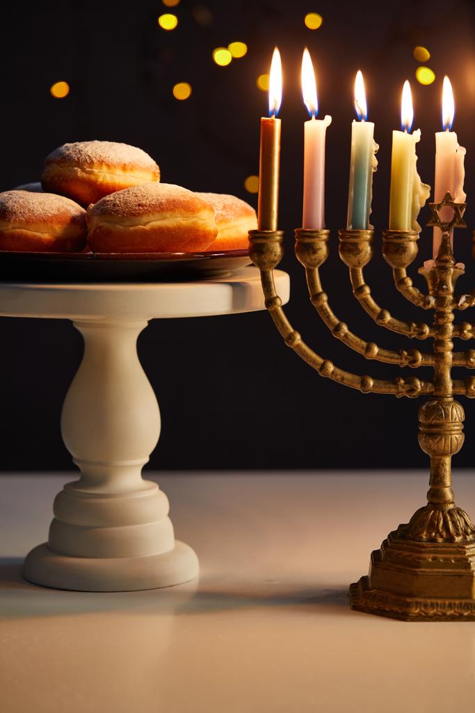 delicious doughnuts on stand near glowing candles in menorah on black background with bokeh lights on Hanukkah - Photo, Image