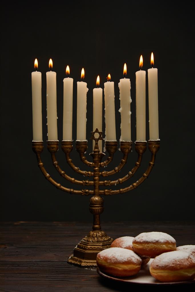 pyszne pączki w pobliżu płonących świec w menorah na Chanuka izolowane na czarny - Zdjęcie, obraz