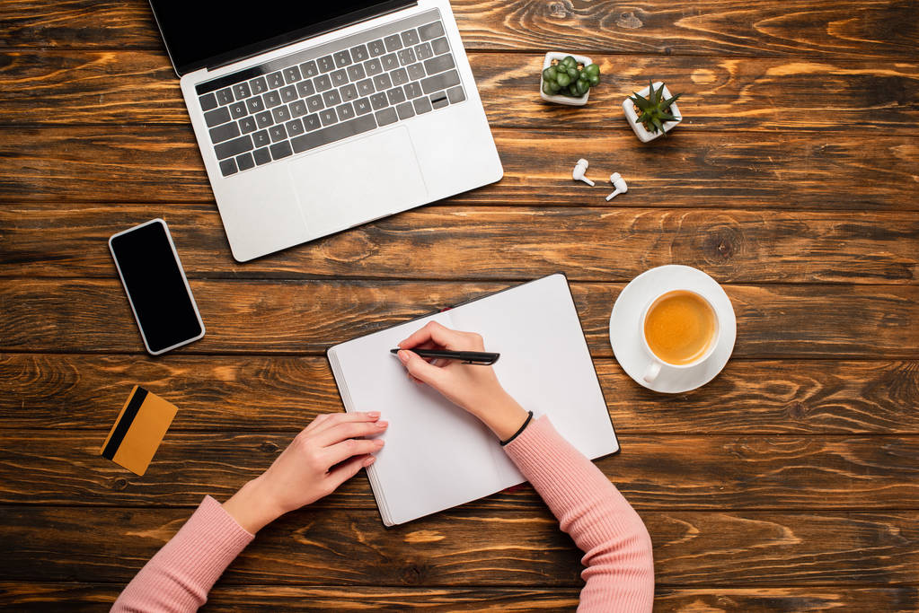 przycięty widok businesswoman pisanie w notebooku w pobliżu laptopa, smartfona, filiżanki kawy, karty kredytowej, słuchawki bezprzewodowe i kalkulator na drewnianym biurku - Zdjęcie, obraz