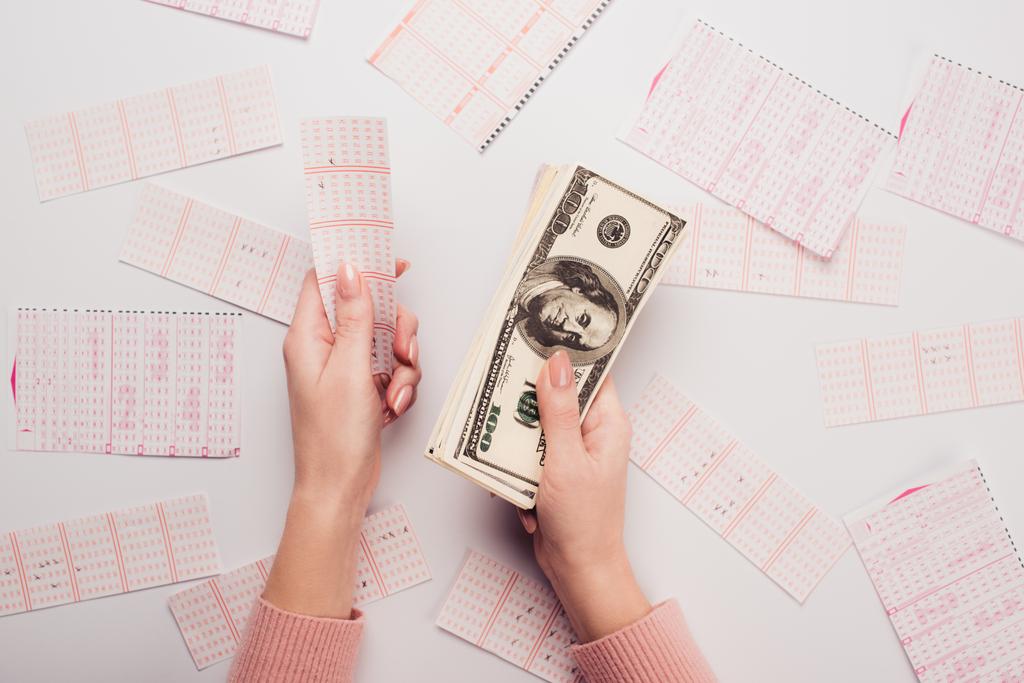 oříznutý pohled na ženu držící dolarové bankovky a los v blízkosti roztroušené loterie karty na bílém stole - Fotografie, Obrázek