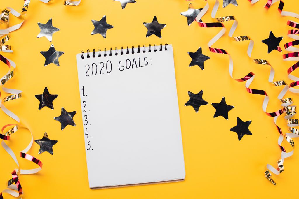 notebook with 2020 goals with empty numbered points near decorative, shiny stars and serpentine on yellow surface - Photo, Image