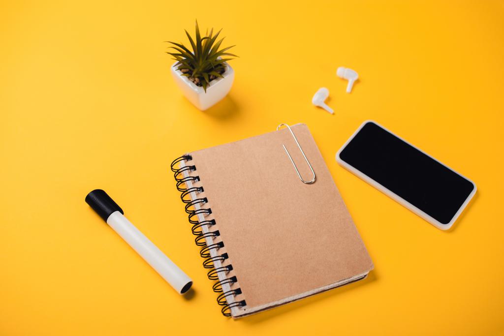 notebook κοντά smartphone με κενή οθόνη, ασύρματα ακουστικά, γλάστρα και στυλό τσόχινη άκρη στο κίτρινο γραφείο - Φωτογραφία, εικόνα