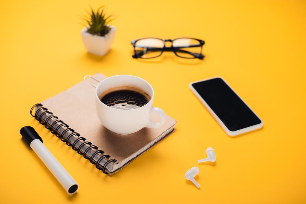 tazza di caffè su notebook vicino smartphone, auricolari wireless, occhiali, pianta in vaso e pennarello sulla scrivania gialla
 - Foto, immagini