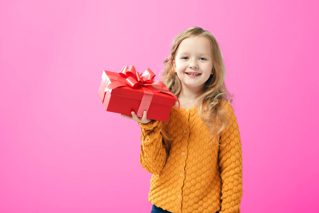 Niña feliz en un suéter de punto cálido sosteniendo una caja con un regalo, aislado sobre fondo rosa. El concepto de dar y recibir regalos
 - Foto, Imagen