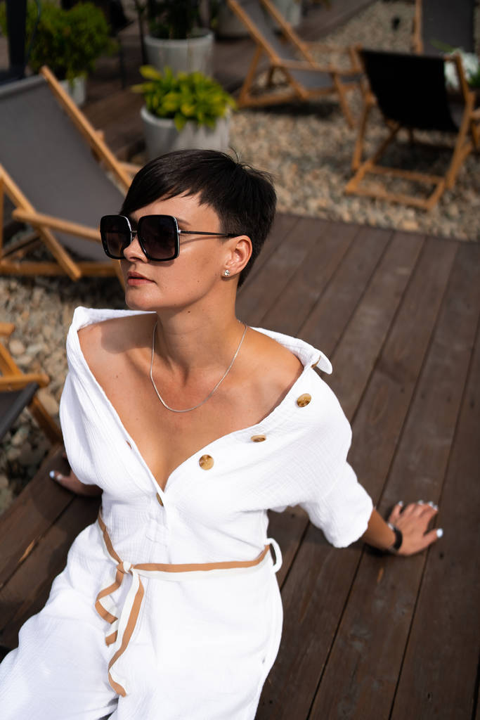 Όμορφη γυναίκα με κοντά καστανά μαλλιά σε λευκά ρούχα και γυαλιά ηλίου. Φωτογραφία δρόμου μόδας. Μόδα μοντέλο κάθεται και να χαλαρώσετε σε καφέ φόντο ξύλο. κατάστημα γυαλιών - Φωτογραφία, εικόνα
