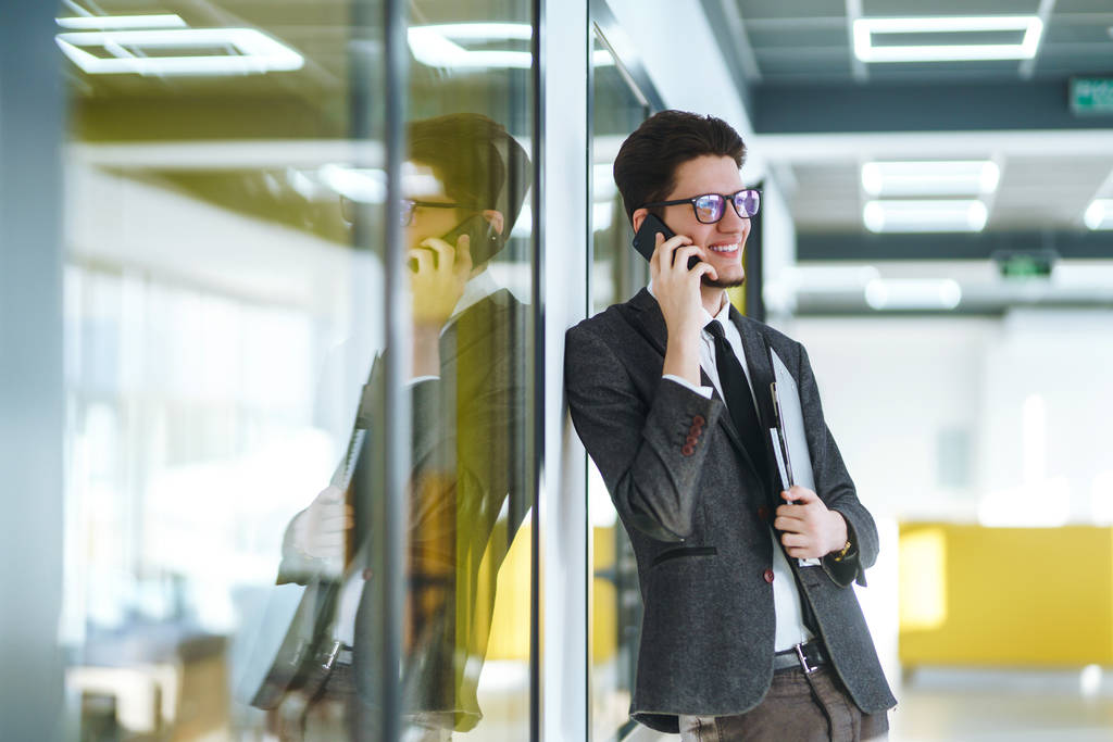 Fiatal üzletember szemüvegben, mobilon beszél a modern irodában. Stílusos férfi öltönyben, mappával és telefonnal. Kommunikációs elv. - Fotó, kép