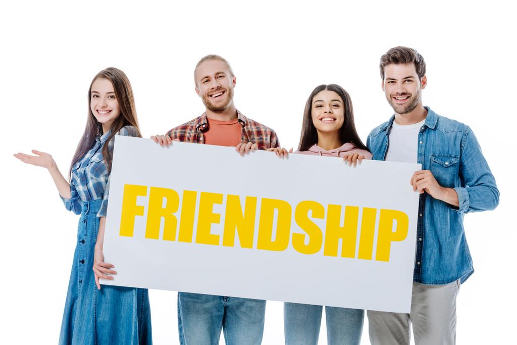 szczęśliwy wielokulturowych przyjaciół trzymających tabliczkę z żółtą przyjaźń liternictwo izolowane na białym - Zdjęcie, obraz