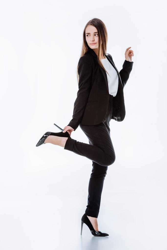 Ganzkörperansicht einer eleganten Geschäftsfrau im Anzug, die mit erhobenem Bein posiert und isoliert auf weißem Grund wegschaut - Foto, Bild