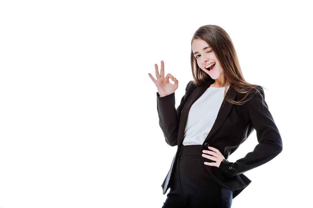 Augenzwinkernde und lächelnde Geschäftsfrau im Anzug zeigt Ok-Zeichen auf weißem Grund - Foto, Bild
