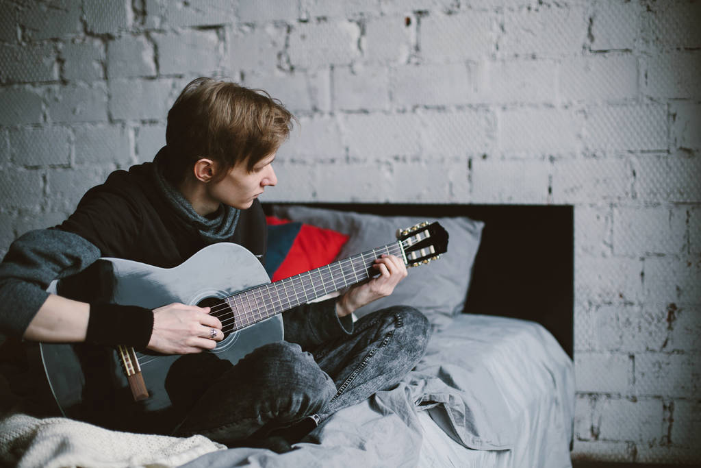 junger männlicher Gitarrist sitzt auf dem Bett und spielt Gitarre. weiße Ziegelwand auf Hintergrund - Foto, Bild