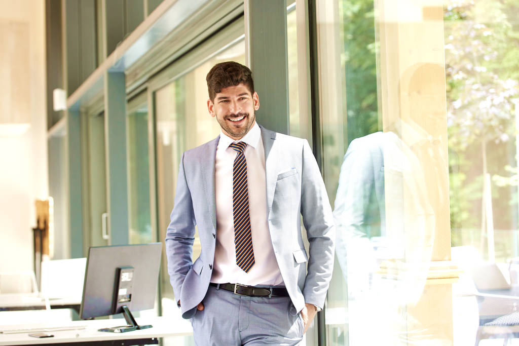 Portret van een zelfverzekerde jonge zakenman in pak en stropdas terwijl hij ontspannen in het raam van het kantoor.  - Foto, afbeelding