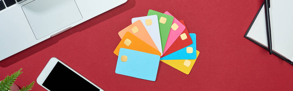 bovenaanzicht van veelkleurige lege creditcards op rode achtergrond met smartphone, laptop, oortjes en koffie, panoramisch shot - Foto, afbeelding