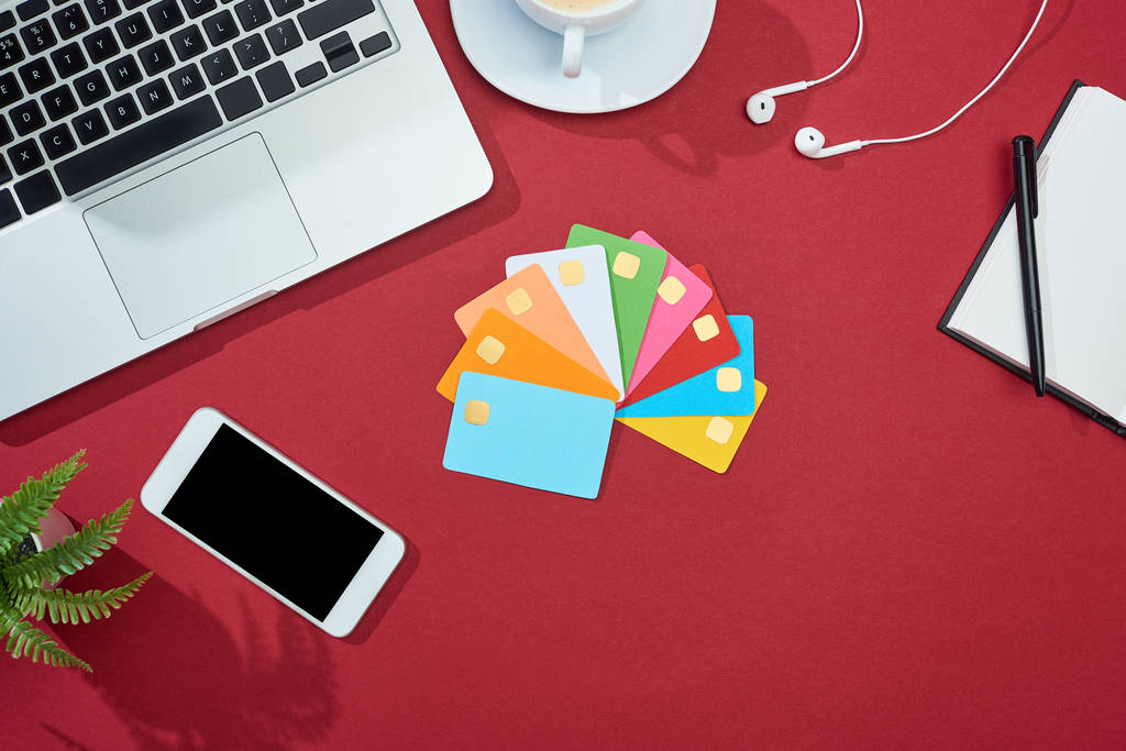 pohled na pestrobarevné prázdné kreditní karty na červeném pozadí s chytrým telefonem, notebookem, sluchátky a kávou - Fotografie, Obrázek