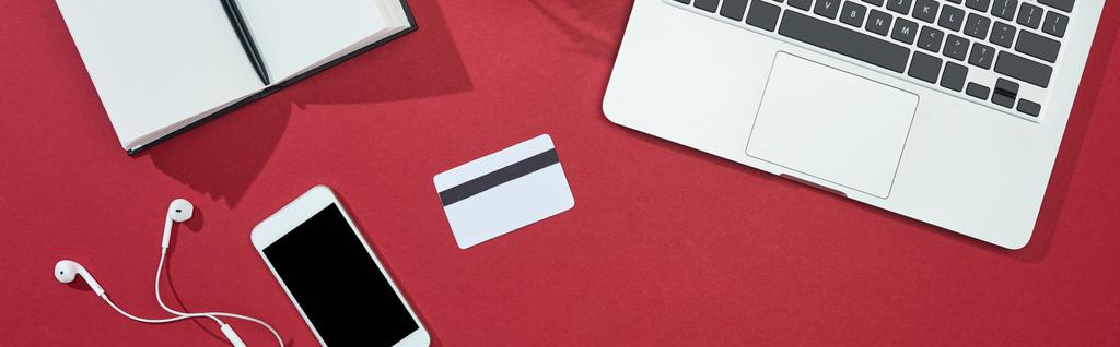 κορυφαία άποψη της πιστωτικής κάρτας σε κόκκινο φόντο με smartphone, laptop, ακουστικά, notebook, πανοραμική λήψη - Φωτογραφία, εικόνα