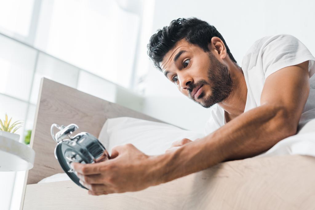 low angle view of shocked bi-racial man holding alarm clock in morning  - Photo, Image