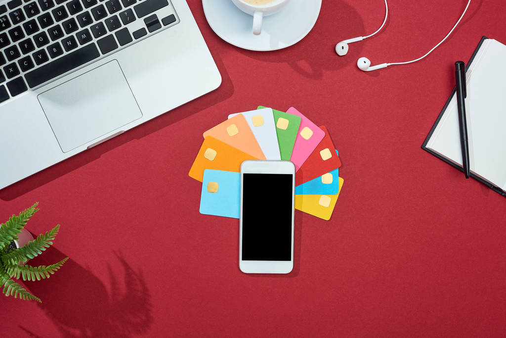 Ansicht von bunten leeren Kreditkarten auf rotem Hintergrund mit Smartphone, Laptop, Kopfhörer und Kaffee - Foto, Bild