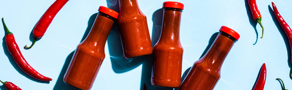 Top view of bottles with chili sauce beside chili peppers on blue surface, panoramic shot - Photo, Image