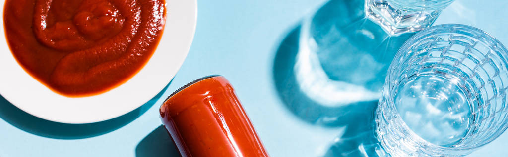 Vista superior de la salsa de tomate casera en plato y botella junto a vasos de agua sobre fondo azul, plano panorámico
 - Foto, Imagen