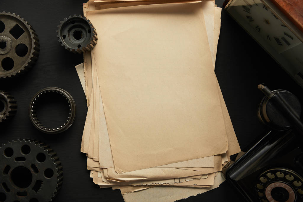 bovenaanzicht van verouderde tandwielen en vintage blanco papier in de buurt van vintage klok op zwart oppervlak - Foto, afbeelding