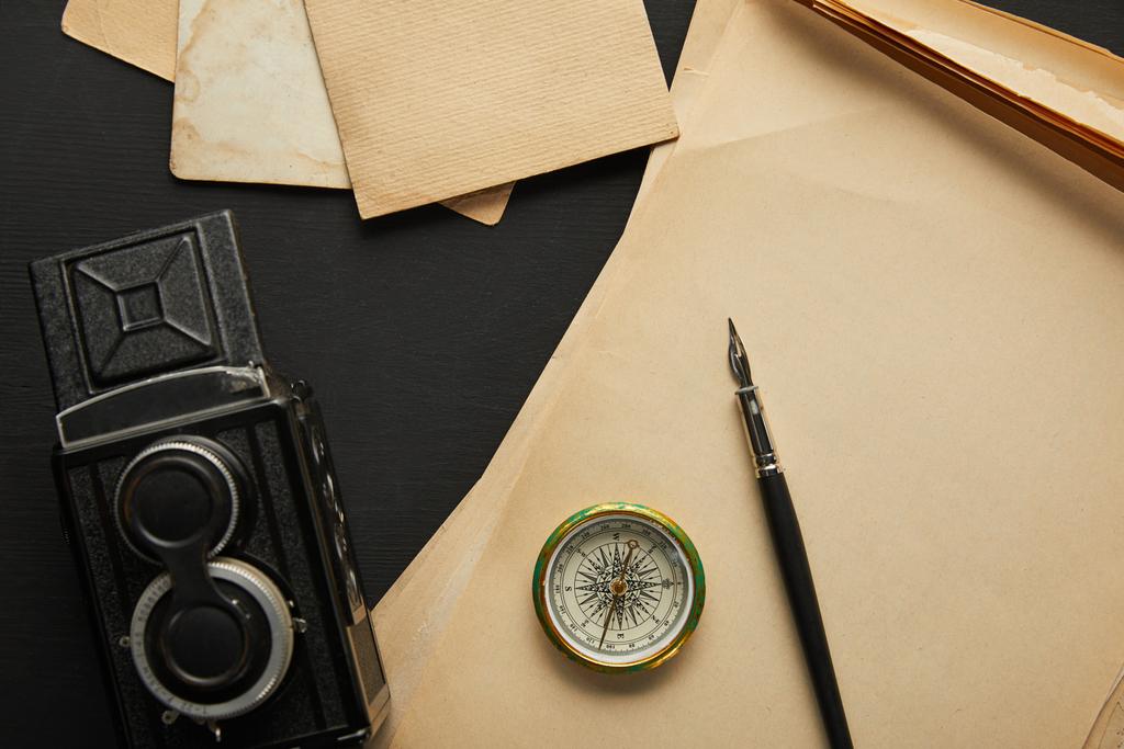 Draufsicht auf alte Kamera, Papier, Füllfederhalter, Kompass auf schwarzem Hintergrund - Foto, Bild